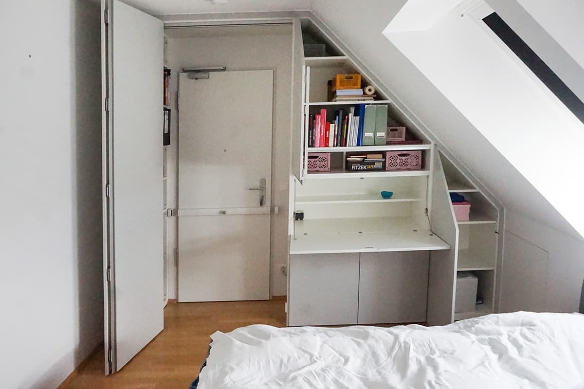 Derselbe Einbauschrank mit geöffneten Türen: Regalfächer mit Ordnern, eine heruntergeklappte Arbeitsfläche und schmale Fächer, die der Dachschräge folgen.