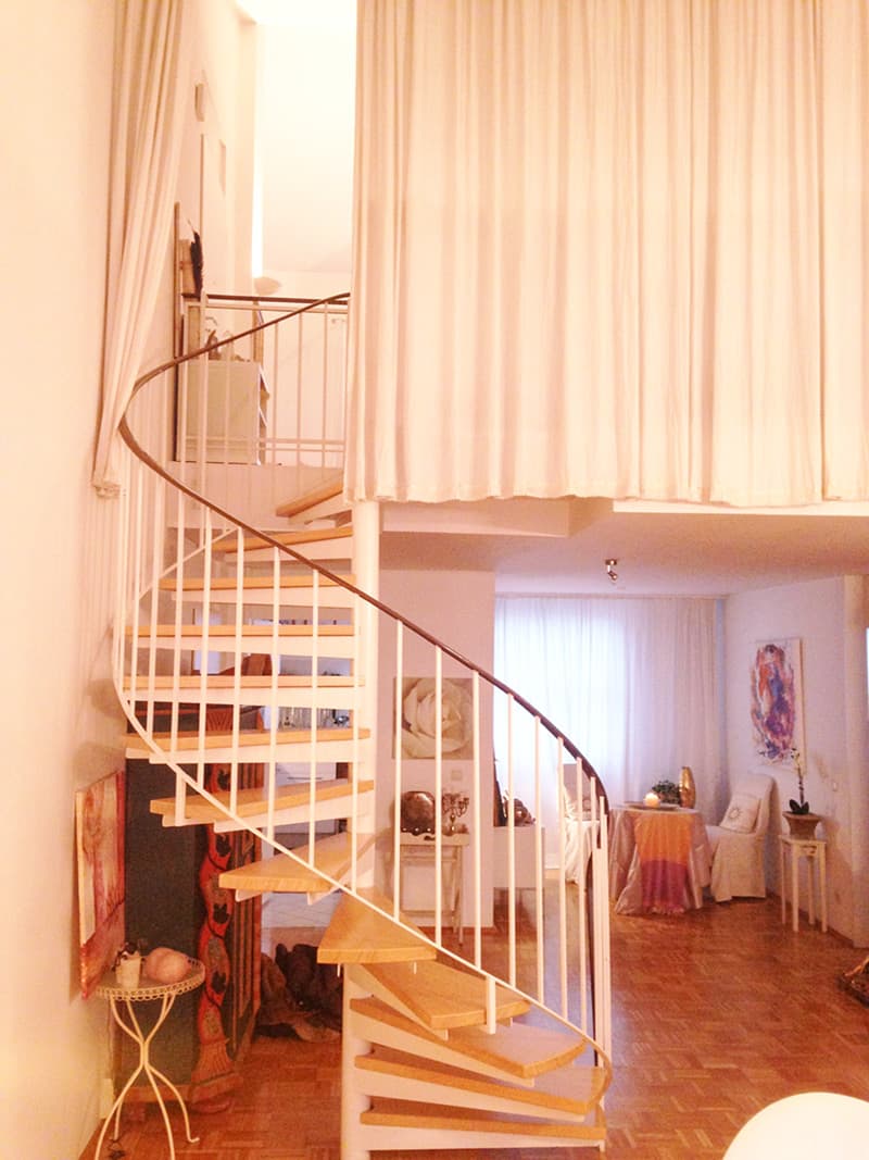 Wendeltreppe mit Stufen aus Holz und weißem Geländer, die in ein offenes Obergeschoss führt.